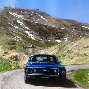 Ford Mustang blu classica su strada di montagna con pendii erbosi e neve residua sullo sfondo
