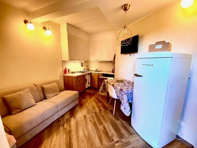 Monolocale con divano beige, angolo cottura in legno e frigorifero bianco con pavimento effetto parquet