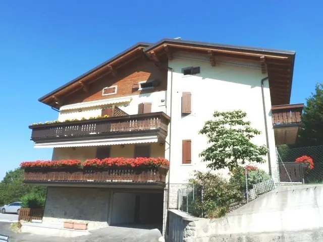 Casa alpina bianca con balconi in legno decorati da fiori rossi, tetto spiovente e cielo azzurro